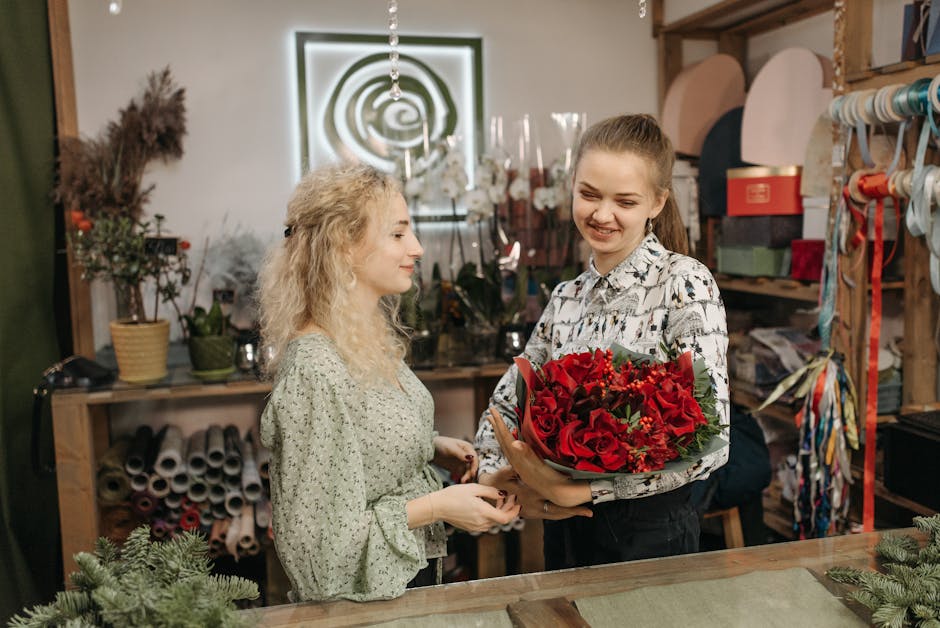 A florist at a floral shop, holding a vibrant bouquet of red roses, gerbera daisies, and green foliage, wrapped in green paper with a ribbon. The florist, a young woman with long hair, is smiling and 