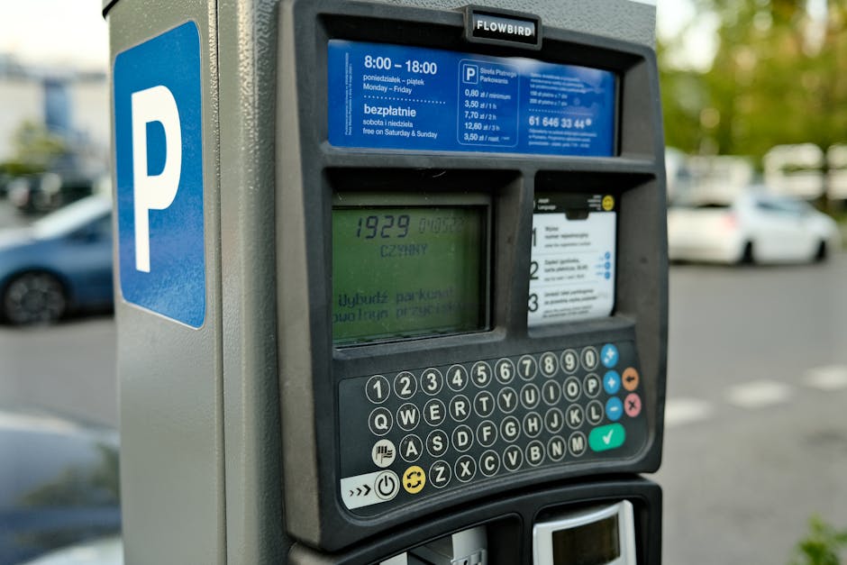 A close-up view of a modern parking meter with a digital screen displaying the time remaining, which shows 19:29, in an outdoor parking lot. The meter has a keypad with black buttons, some marked with