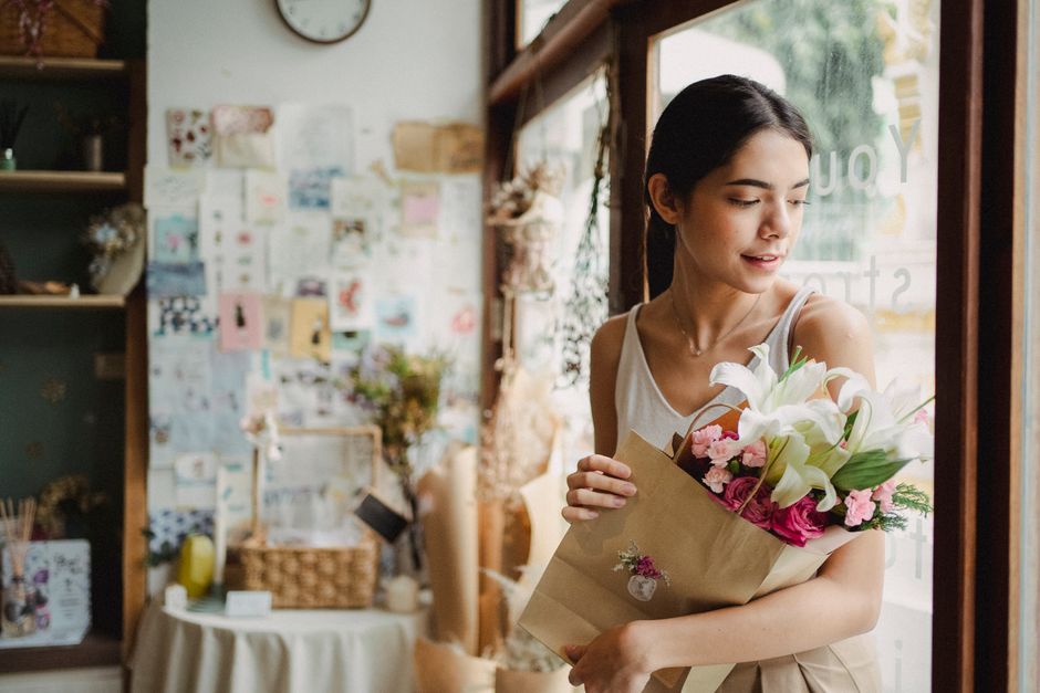 A young woman with dark hair, wearing a white sleeveless top, is holding a bouquet of fresh flowers wrapped in brown paper inside a cozy flower shop. The bouquet features white lilies with large petal