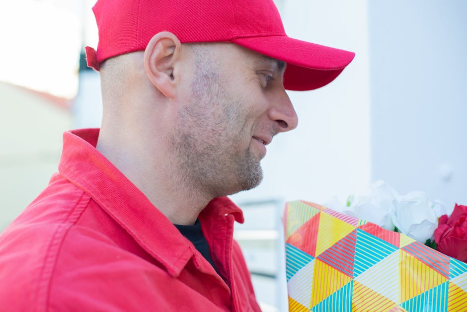 Close-up of a delivery person wearing a red cap and red jacket holding a colorful, geometrically patterned gift bag filled with fresh, vibrant flowers, including red roses and white blooms, in a well-