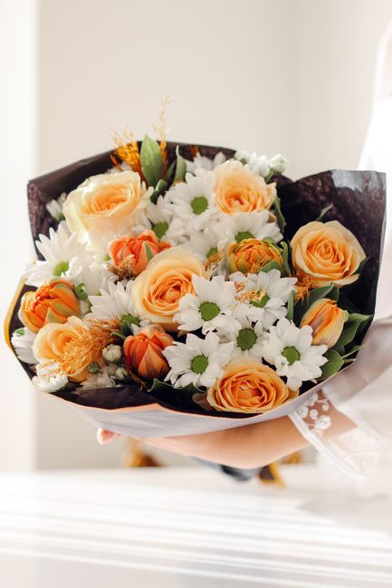 A hand holding a beautifully arranged bouquet of fresh flowers, featuring peach-colored roses, white daisies with yellow centers, and small orange tulips, all wrapped in dark purple and white tissue p