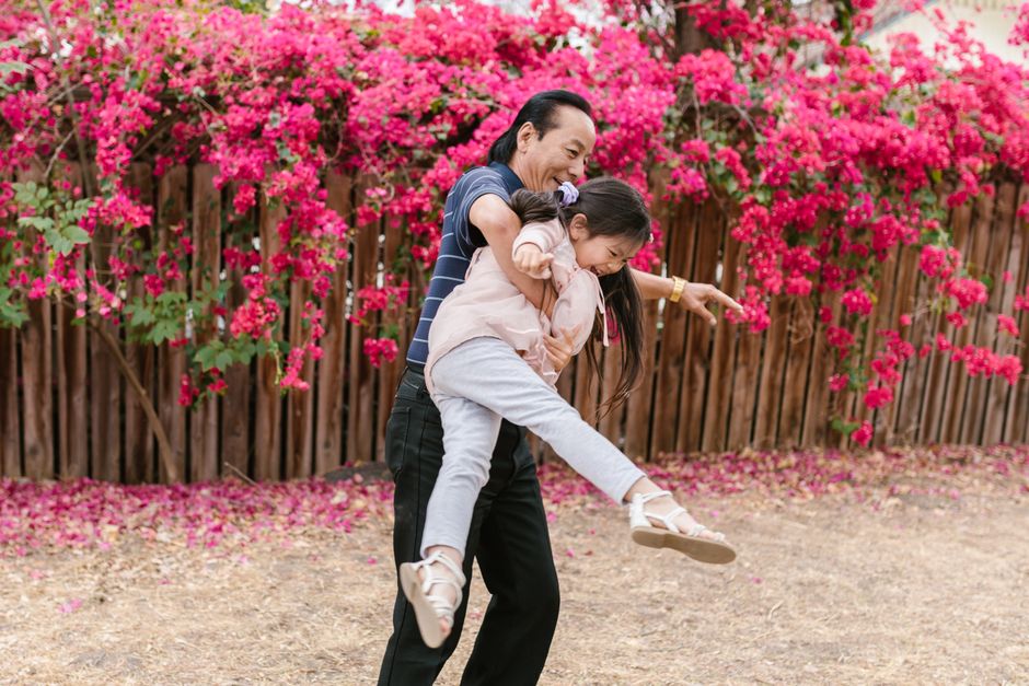 A man with dark hair and a striped navy T-shirt is smiling and holding a young girl with long dark hair, dressed in a pink jacket and light gray pants, in a playful lift outdoors. The girl is extendin