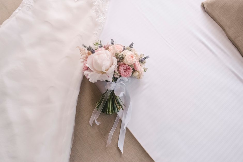 A delicate bouquet of fresh pink and white flowers, including peonies and roses, arranged with small sprigs of lavender and grey foliage. The bouquet is wrapped in white tissue paper and tied with a s