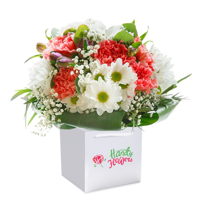 Presented upright in a square white gift bag with sturdy ribbon handles, this bouquet showcases the careful touch of an experienced florist: coral-pink and peach carnations gather into a plush, ruffled mass, their layers catching soft light across each velvety petal. Dotted throughout are snow-white daisy chrysanthemums with lime-green centres that add a crisp, cheerful punctuation to the warmer tones. Wisps of baby's breath (Gypsophila) weave through the blooms, creating a delicate, cloud-like texture that softens the overall silhouette. A few closed buds with a purple tinge peek from one side, introducing a subtle depth and the anticipation of unfolding colour-likely small alstroemeria or similar buds. Broad, glossy leaves fan around the base, forming a rich green collar that stabilises the arrangement and contrasts beautifully with the pastel and vivid florals. Photographed against a clean white background, each element feels distinct: the carnations' velvety softness, the daisies' crisp brightness, and the greenery's glossy sheen. This composition reads as an elegant, ready-to-gift choice ideal for marking joyful moments: birthdays, a heartfelt thank-you, or a simple pick-me-up for someone living near Emerson Park Village or catching the train at Emerson Park station. For customers seeking local, fresh delivery, Flowers Emerson Park can prepare and present this arrangement with the same attentive craftsmanship visible here.