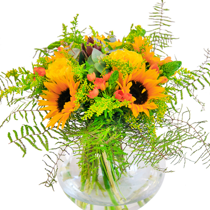 Graceful, abundant, and glowing in a spectrum of yellows and oranges, this seasonal bouquet embodies the spirit of Emerson Park's sunlit gardens. Towering sunflowers command attention at its center, their broad, dark hearts ringed by generous petals whose color recalls autumn leaves scattered along Slewins Lane. Velvet yellow garden roses spiral open, each layer revealing a new dimension of texture and softness that speaks to both elegance and comfort. Scattered throughout, glossy red Hypericum berries bring an unexpected playfulness, their bold hues enlivening both the arrangement and the senses. A whisper of feathery fern, interlaced with sprigs of goldenrod and slender, silvery eucalyptus, provide a nuanced green backdrop-cool, fresh, and reminiscent of Emerson Park's abundant greenery. Every stem's architecture is clearly revealed through the transparent, rounded glass vase; water glimmers below, enhancing the bouquet's radiant tones. The air is filled with a subtle blend of floral and green notes, inviting memories of crisp walks through Harrow Lodge Park under late summer skies. More than just a display, this bouquet is crafted to mark joyful milestones-a birthday, anniversary, or a simple act of kindness-imbued with the warmth and artistry of Flowers Emerson Park.