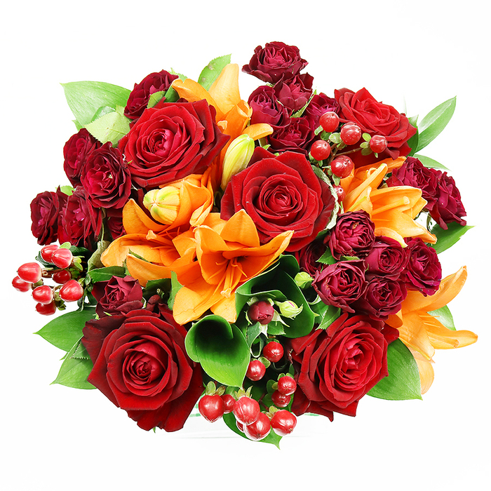 Captured from above against a spotless white field, this opulent, hand-tied bouquet is a harmonious chorus of deep reds, bright oranges, and verdant greens that feels both sumptuous and quietly joyful. The composition is tightly arranged in a circular silhouette: prominent, fully opened deep red roses form the anchoring centre, their velvety petals curling to reveal rich, layered centres that read as timeless romance. Sprung between those large blooms are orange Asiatic lilies, their trumpet shapes and delicate stamens contributing sculptural contrast; some are in full exuberance while others remain in bud, suggesting a bouquet that will continue to develop over days. Nestled among these are dark red spray roses-clusters of miniature roses whose dense, velvety heads add shadow and jewel-tone complexity. Glossy hypericum berries, small and round, appear like bright punctuation marks across the surface, catching highlights and offering a playful texture against the soft petals. Generous swathes of green foliage-likely ruscus or salal-frame the flowers, providing cool relief and structure that lifts and separates the blooms so each colour reads distinctly. The lighting is soft and even, highlighting petal texture without harsh shadows, and the overall mood is celebratory and warm: an autumnal yet timeless arrangement suited to anniversaries, declarations of love, or as a thoughtful house-warming for someone moving into a flat near Emerson Park or a quiet home by Raphael Park. This bouquet pairs well with candlelit evenings and the comforting hush of a neighbourhood evening walk.
