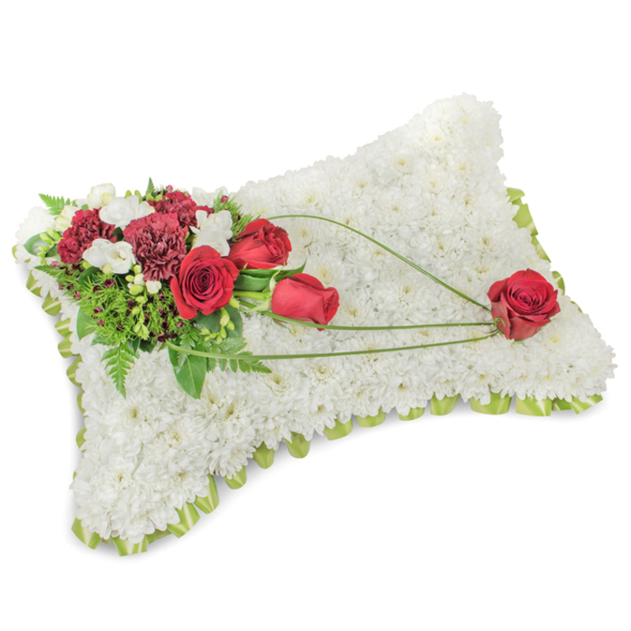 A refined and poignant pillow-style tribute formed from a dense blanket of small white chrysanthemums, each bloom closely set to create an almost textile-like surface that communicates calm, purity and peaceful rest. The rectangular cushion is softly rounded at the corners with a faint outline of pale green ribbon or tender foliage peeking between flower heads to define the edge and lend a subtle freshness. Anchoring the design on the upper left is a richer grouping of deep red roses-some tightly shaped, others opening into full, velvety cups-paired with dark burgundy carnations whose ruffled petals add sculptural weight. Scattered among them are tiny, creamy white accent blooms, likely freesia or small spray chrysanthemums, which bring lightness and a faint fragrance. Layers of green leaves and slender fern fronds form a verdant backdrop for the primary flowers. Extending diagonally from the main spray, three sweeping strands of bright green bear-grass or similar linear foliage trace an elegant arc across the white chrysanthemum surface, guiding the eye to a single, perfectly formed red rose resting near the lower right corner - a considered focal point that evokes memory and affection. The photograph's soft, neutral lighting heightens texture and colour contrast, and the overall arrangement is befitting of services held in neighbourhood chapels or the quiet corners near Emerson Park station, Hornchurch Country Park or local funeral venues. This classic memorial cushion combines thoughtful craftsmanship, comforting hues and dignified sentiment to honour a loved one with reverence.